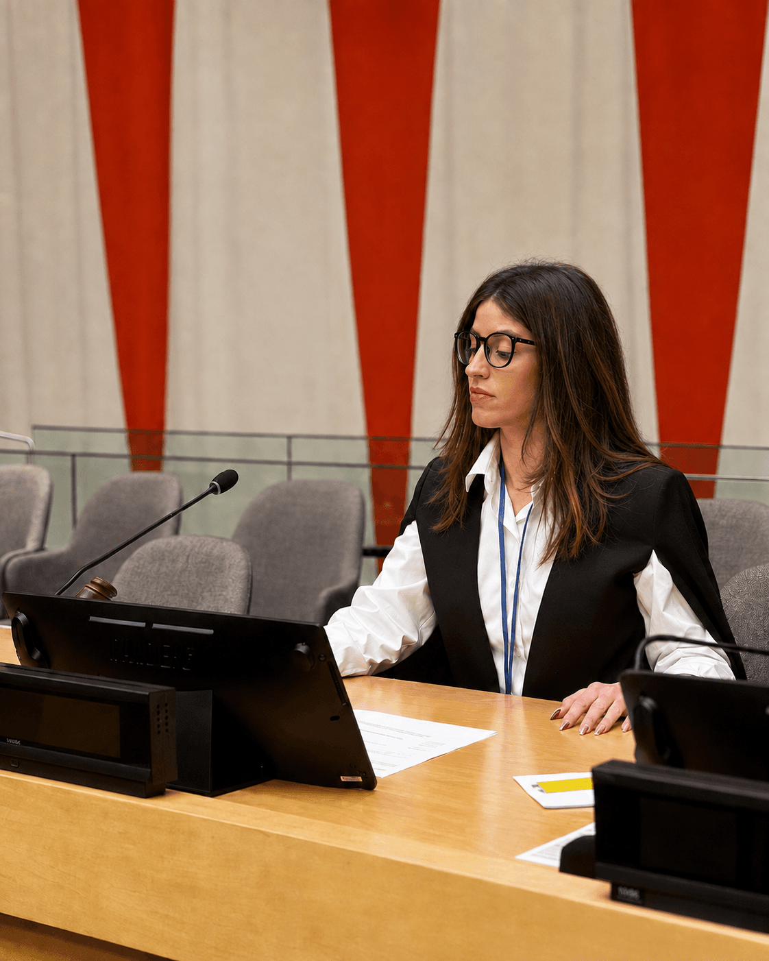 Woman with glasses and black vest sits at a desk with a microphone and papers.